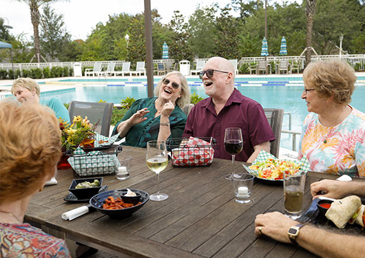 Senior friends laughing and joking while eating lunch pool side.